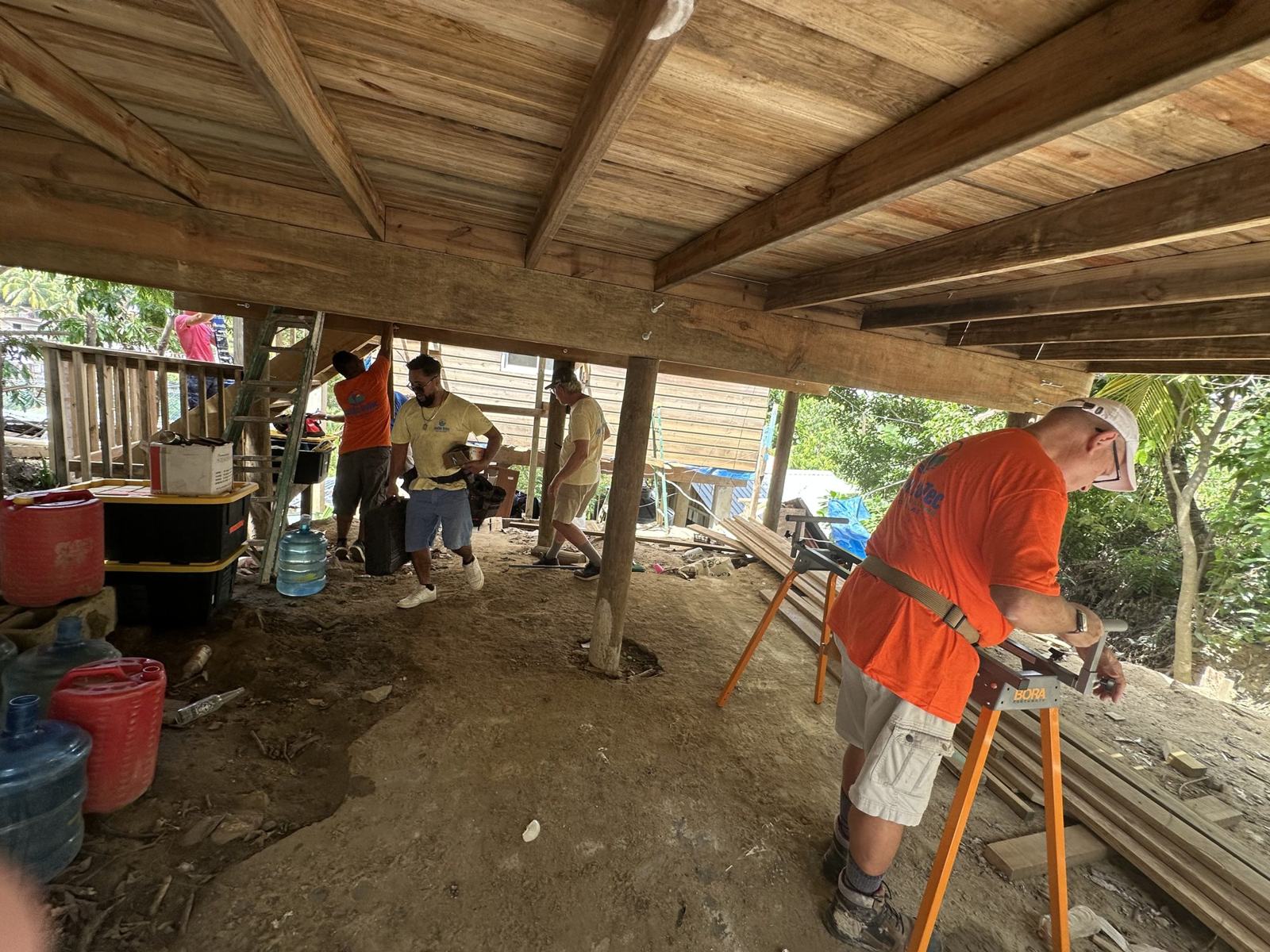 A volunteer cuts lumber on a sawhorse in the shade beneath a raised house
