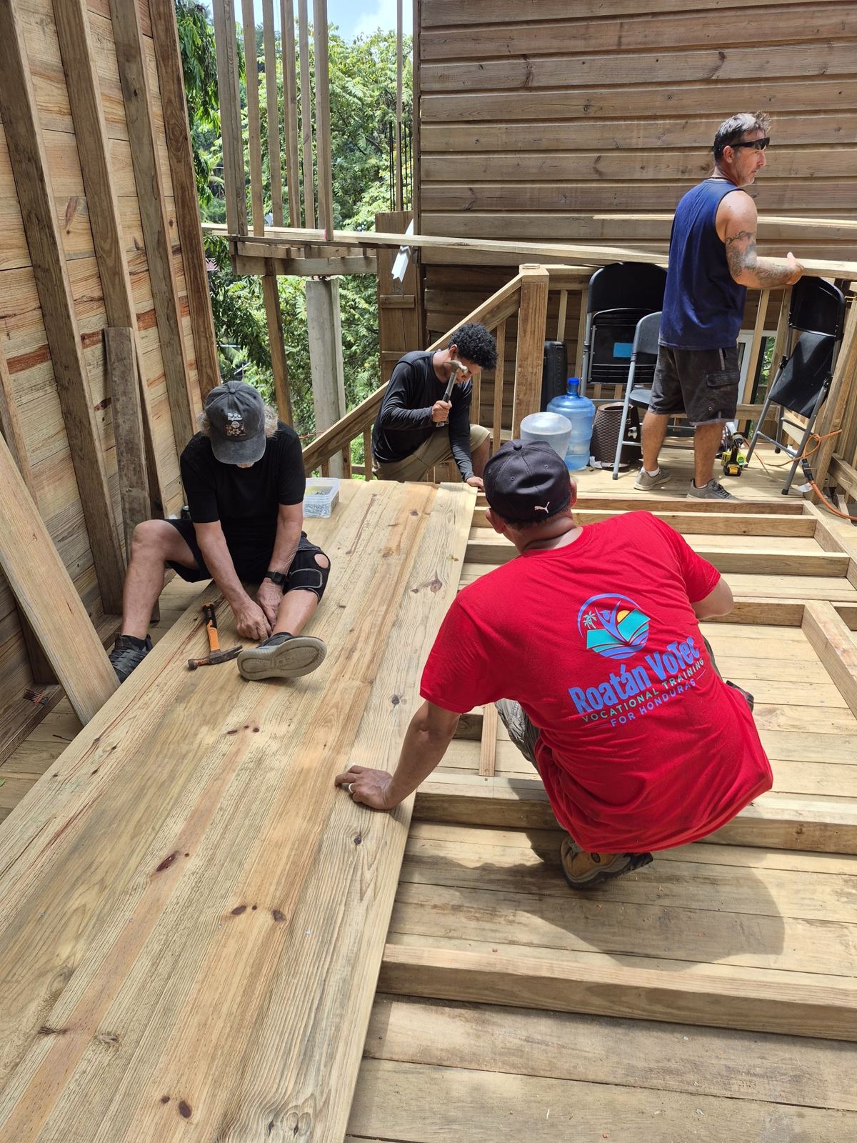 Volunteers and a builder in a red Roatán VoTec shirt nail framing together on the wooden platform