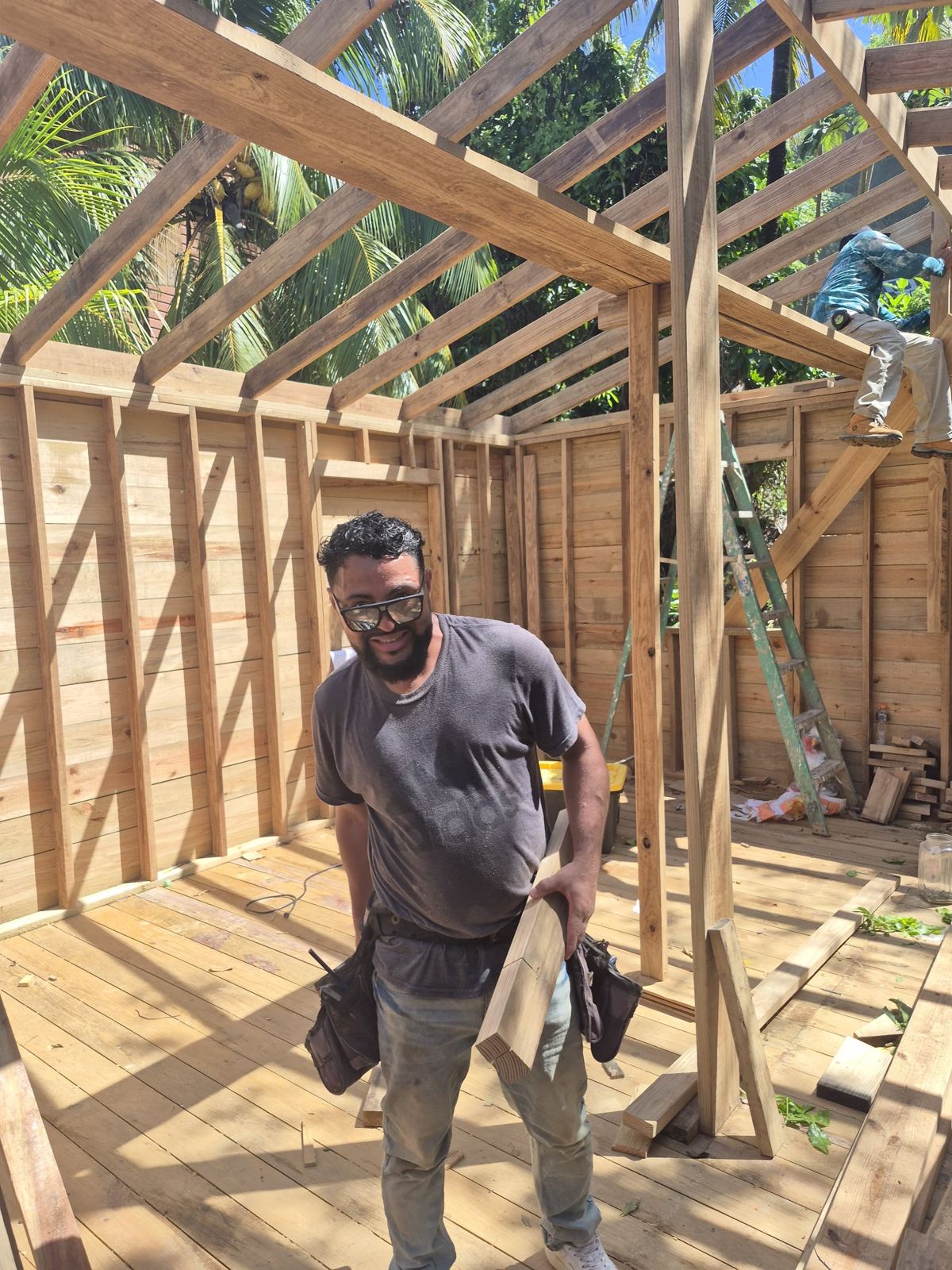 A smiling builder with a tool belt stands inside the framed home under a full set of roof rafters and palm trees