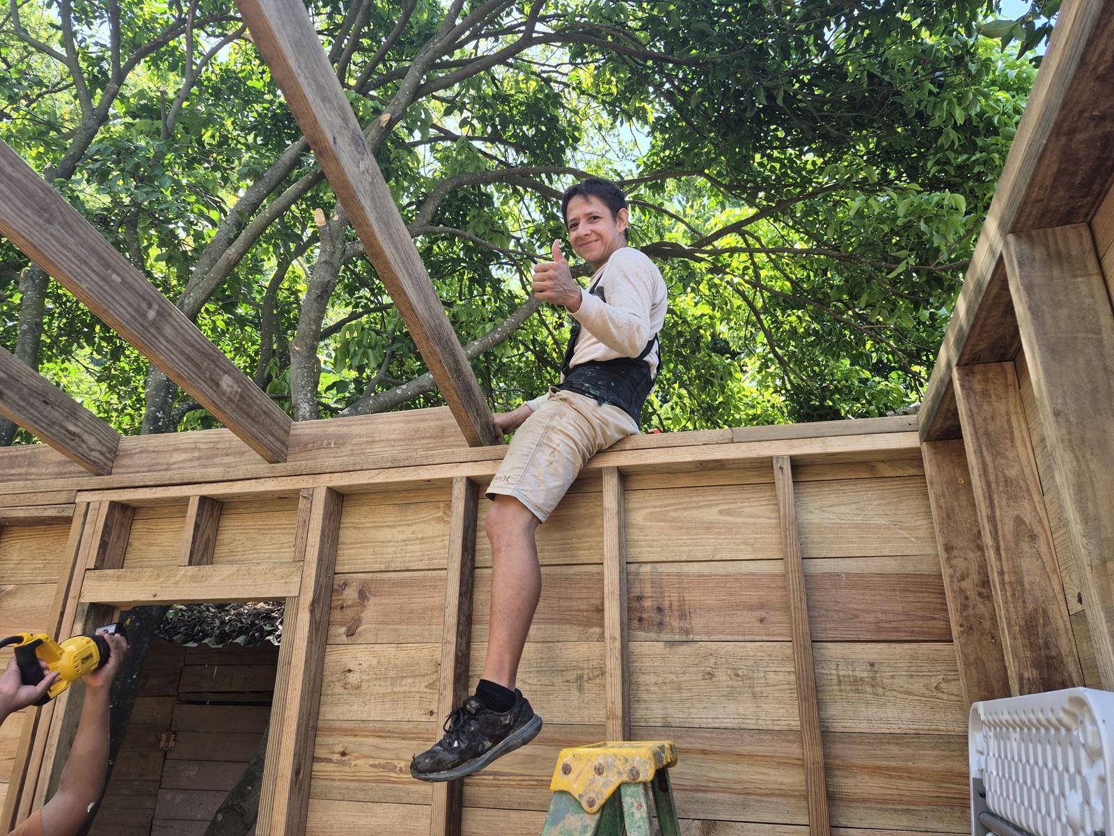A builder sits on top of the framed wall under the rafters and gives a thumbs up