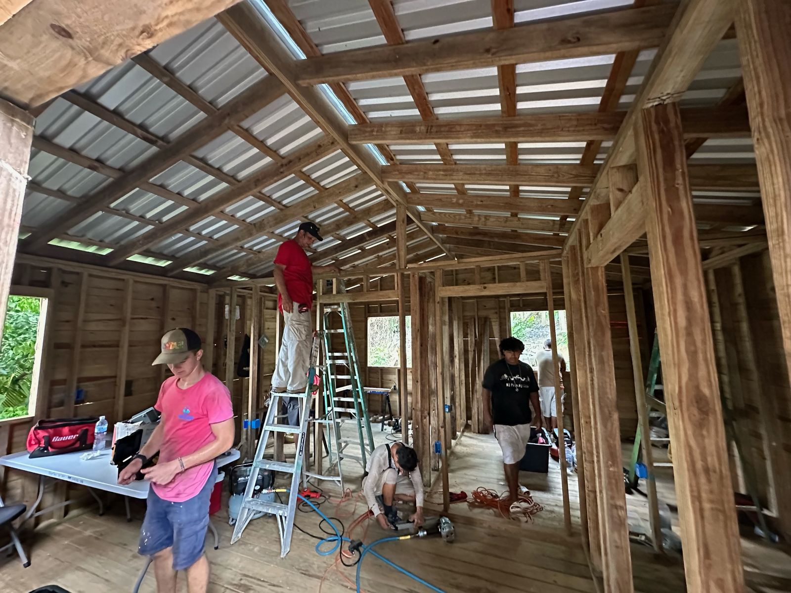 Volunteers work under the new roof: one on a ladder at the top plate, one at the tool table, one drilling at the floor