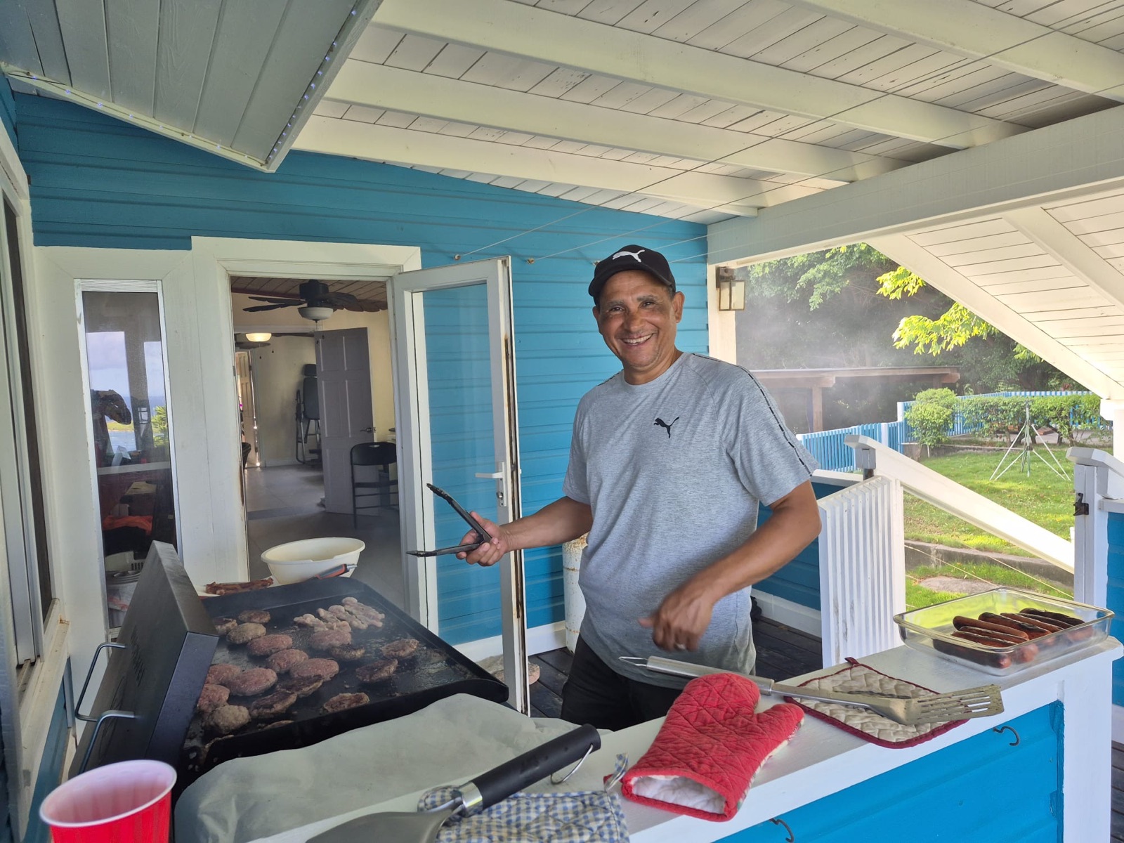 Chef Jose smiles at a flat-top grill loaded with burger patties, a tray of hot dogs beside him on a blue and white porch