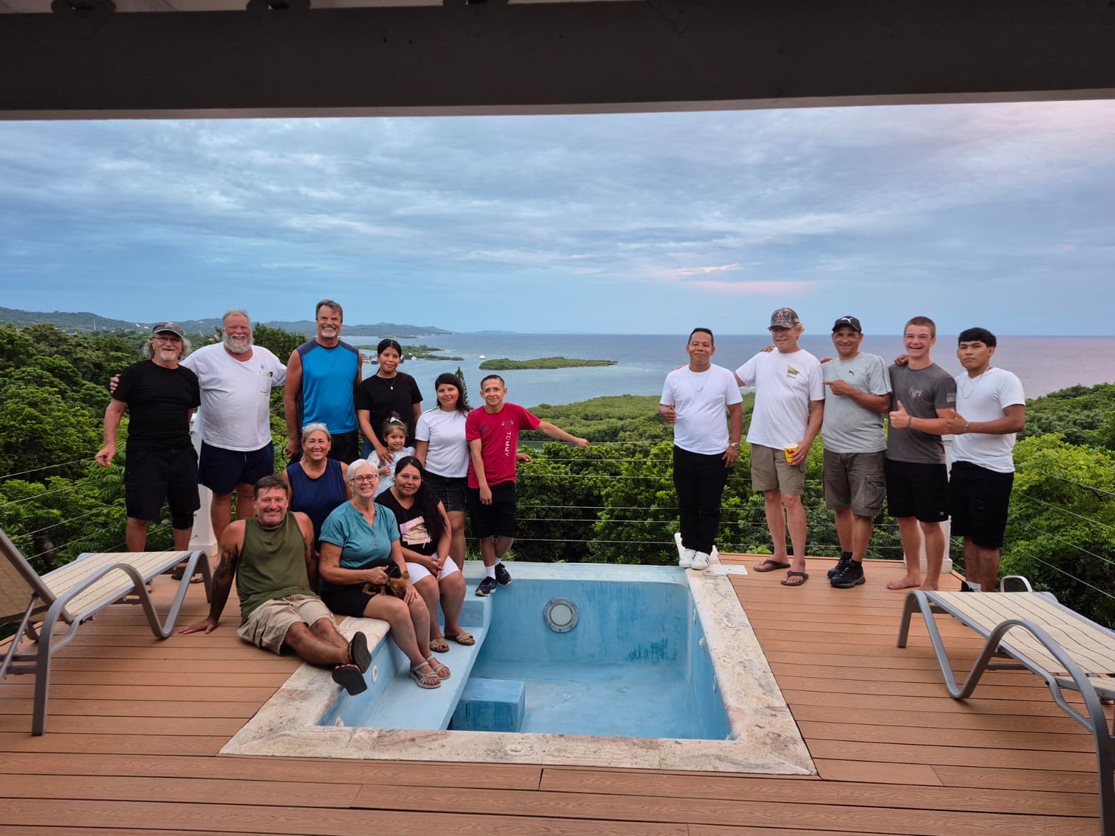 The volunteer team and local builders with their families gathered on a wooden deck above the trees, with the bay and the sea behind them