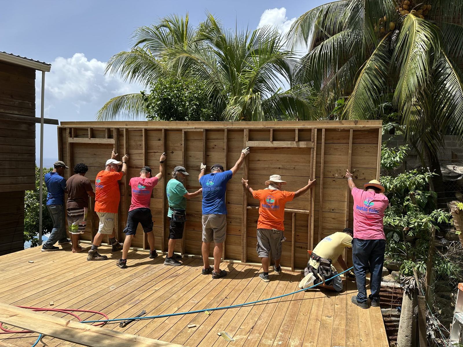 Volunteers and local builders raise the first wall of a new home on a raised wooden platform, palm trees and sea behind