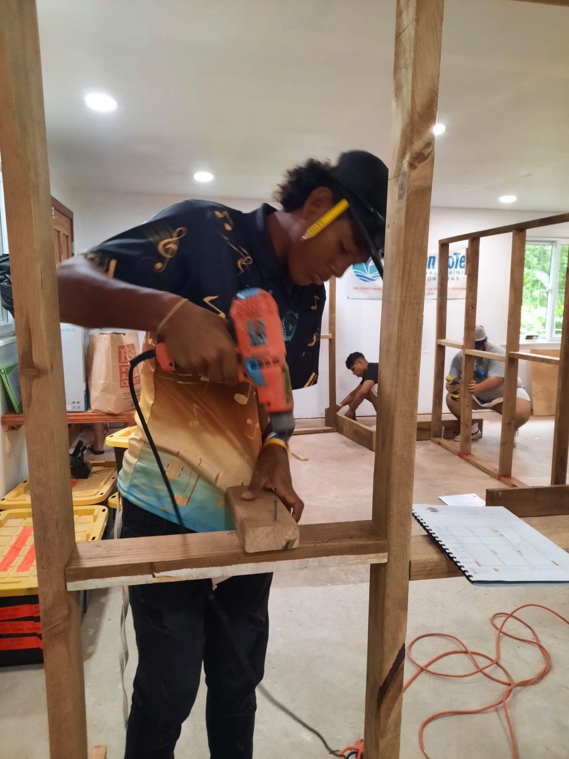 A student with a pencil behind his ear drives a screw into a wooden wall frame with a corded drill while two classmates assemble frames behind him