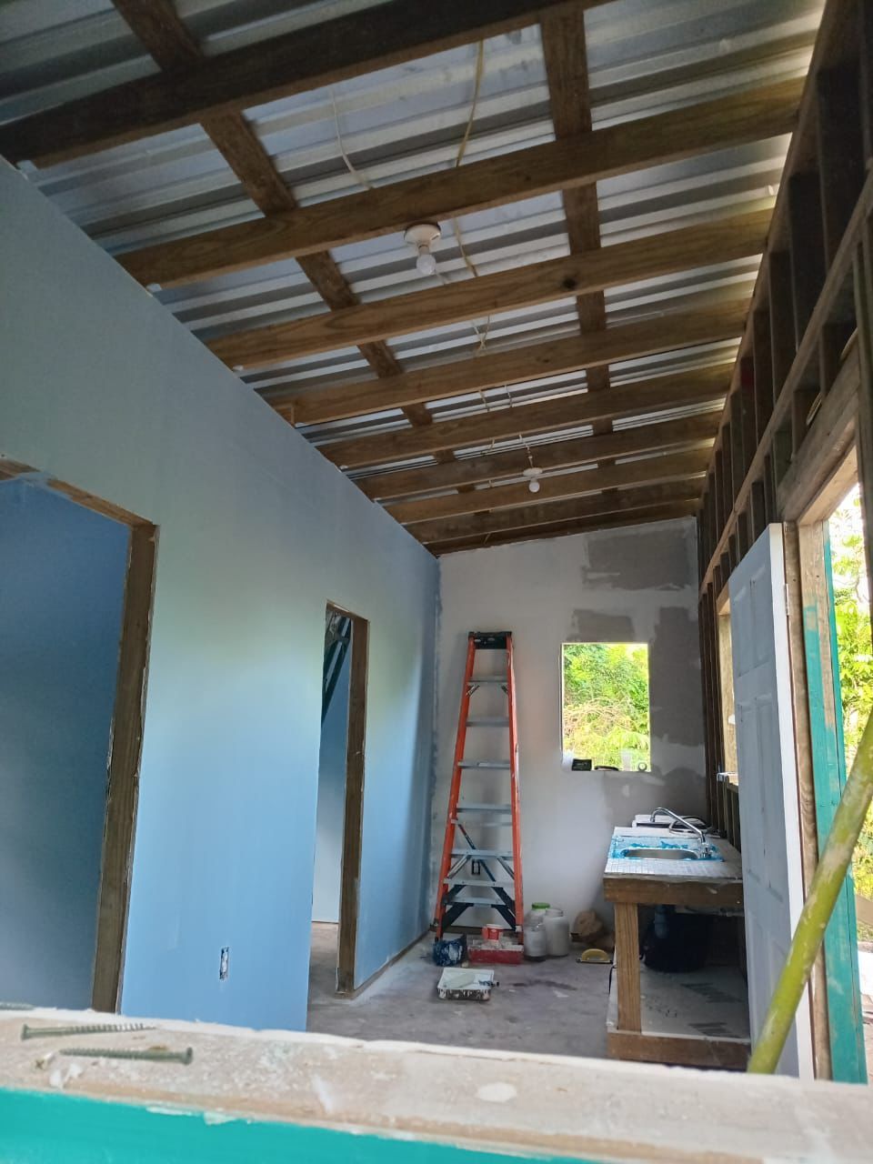 Inside a house under construction: drywall hung and painted light blue, open rafters under a metal roof, a ladder and a sink against the far wall