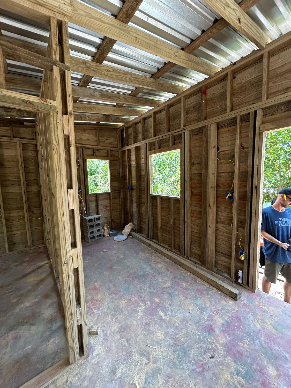 Inside a wood-framed house under a metal roof, with electrical boxes and yellow wire run along the studs and window openings looking out on trees